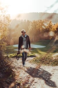 A stylish man in sunglasses, a hoodie, and a jacket carrying a camera, walking on a tree-lined path with autumn leaves and a golf course in the background.